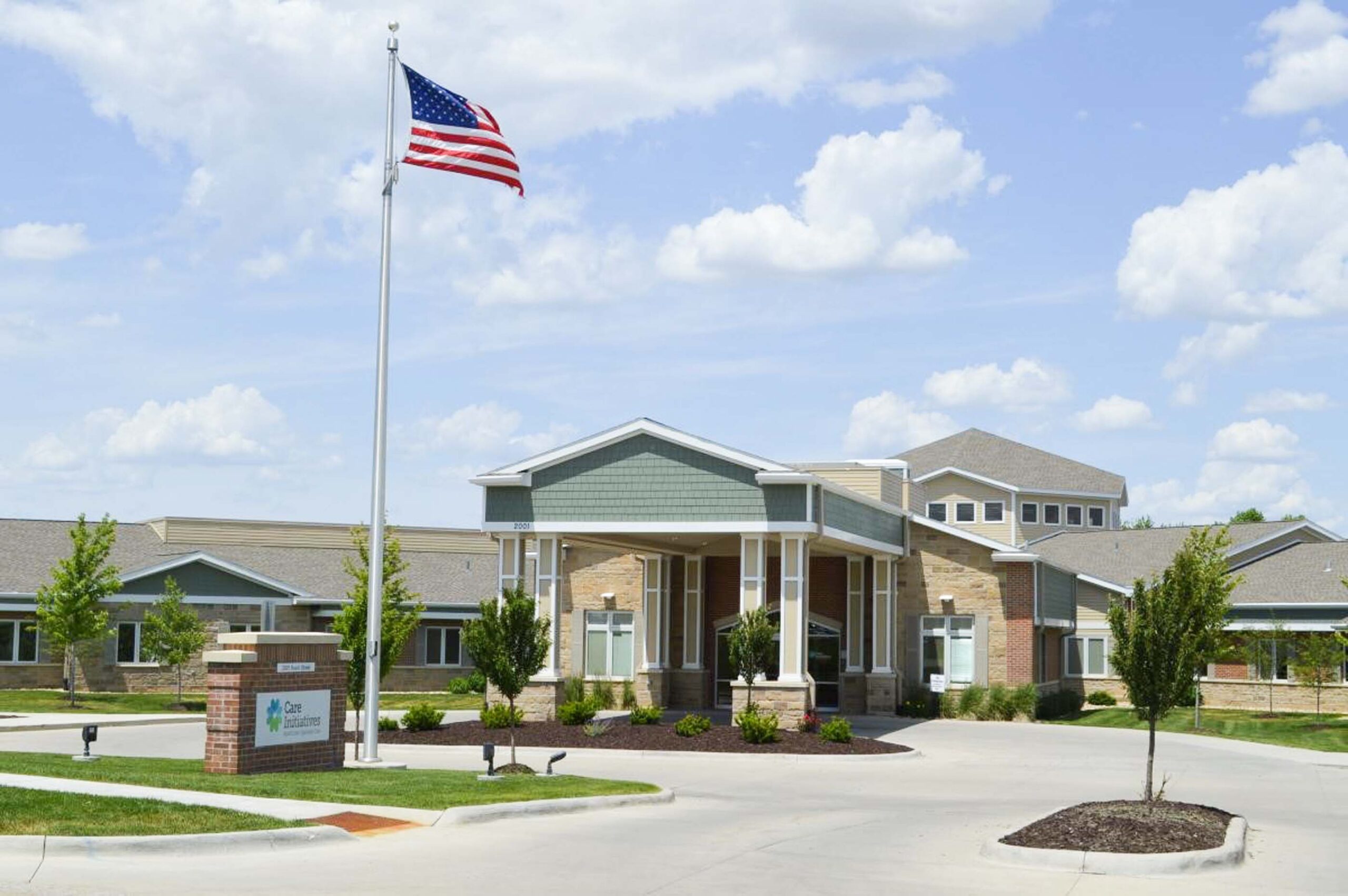 A modern one-story building with a pitched roof is surrounded by a well-manicured lawn and young trees. An American flag flies on a tall pole near the entrance, symbolizing the Care Initiative. A sign with colorful graphics is visible near the driveway under a partly cloudy sky, highlighting the brand name Care Initiative.