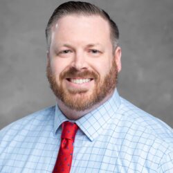 A bearded man smiles at the camera, dressed in a light blue checkered shirt with a red tie against a plain gray backdrop. His confident demeanor hints at his involvement in an important project for Care Initiative.