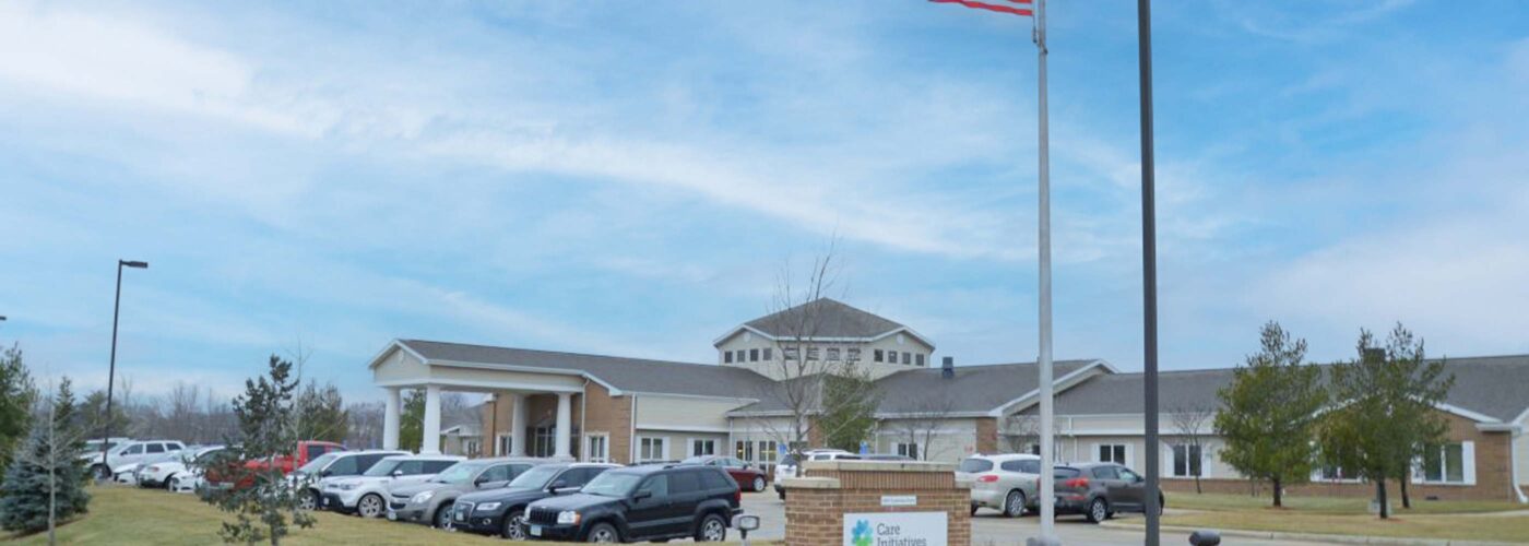 A large building with a sign for Care Initiative is surrounded by a parking lot filled with numerous cars. An American flag flies on a flagpole in the foreground, while trees and a grass lawn extend under a cloudy blue sky.