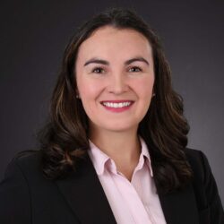 A person with long, wavy dark hair smiles while wearing a black blazer and a pink shirt. The dark gray background enhances the professional portrait atmosphere, embodying a sense of care and initiative in their expression. Care Initiative.