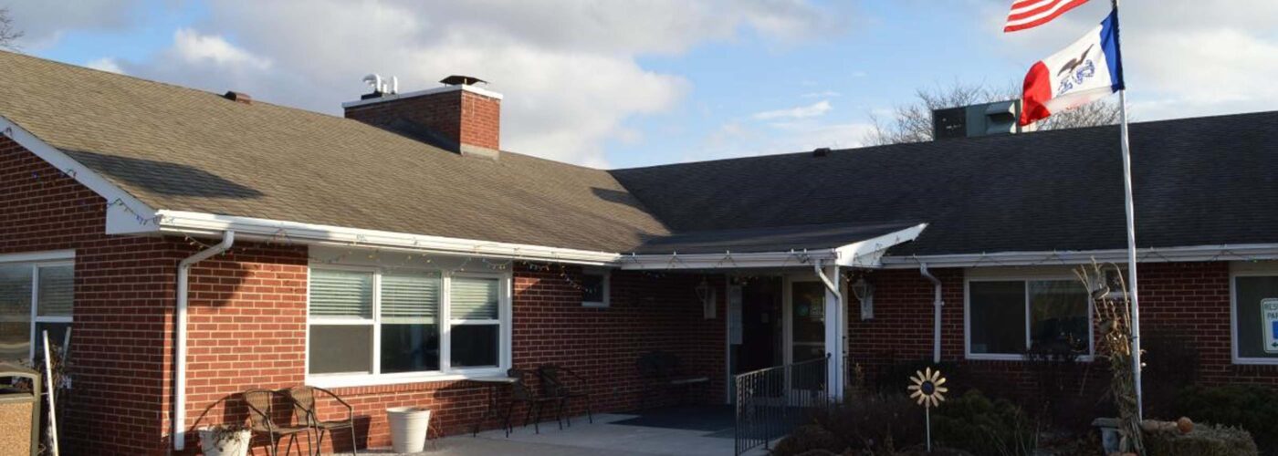 A brick building with a sloped roof displays two flags: the U.S. flag and one adorned with a bird symbol. Emphasizing accessibility, it features an entrance ramp and patio chairs scattered around, all beneath a partly cloudy sky. This reflects Care Initiative's commitment to inclusivity.