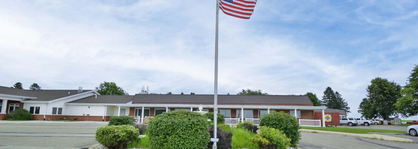 A single American flag stands proudly in the center of a circular garden with bushes, surrounded by a paved driveway. In the background, as part of a broader Care Initiative, is a one-story building with a light-colored exterior under a partly cloudy sky.