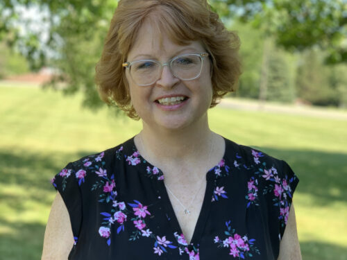 A smiling woman with short red hair and glasses stands outdoors, wearing a black shirt with pink and purple floral patterns. The background features green grass and leafy trees in sunlight. Care Initiative.