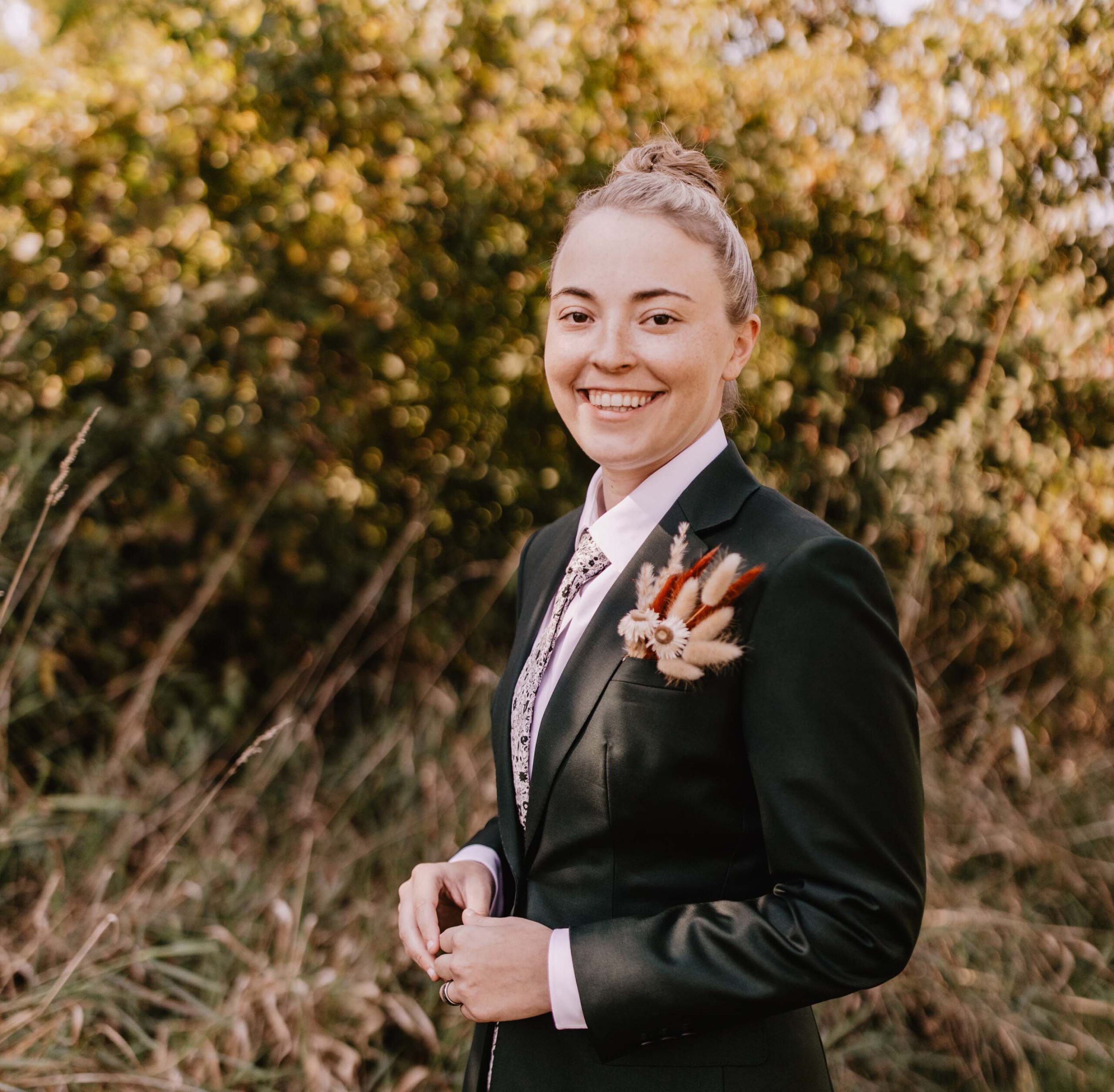 A person in a dark suit and patterned tie stands outdoors, smiling. The suit features a unique brooch with feathers. Lush green foliage and tall grass create a natural setting, evoking a sense of harmony often promoted by Care Initiative.