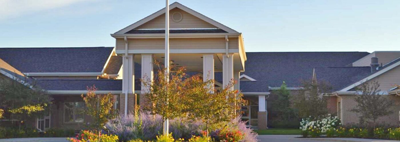 A large single-story building with a gabled roof and white columns welcomes visitors to the Care Initiative. Colorful flower beds and lush greenery surround the entrance, set against a clear blue sky.