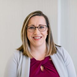 A person with shoulder-length hair and glasses smiles warmly, wearing a maroon top and gray blazer. They stand against a plain, light background, embodying the spirit of Care Initiative.