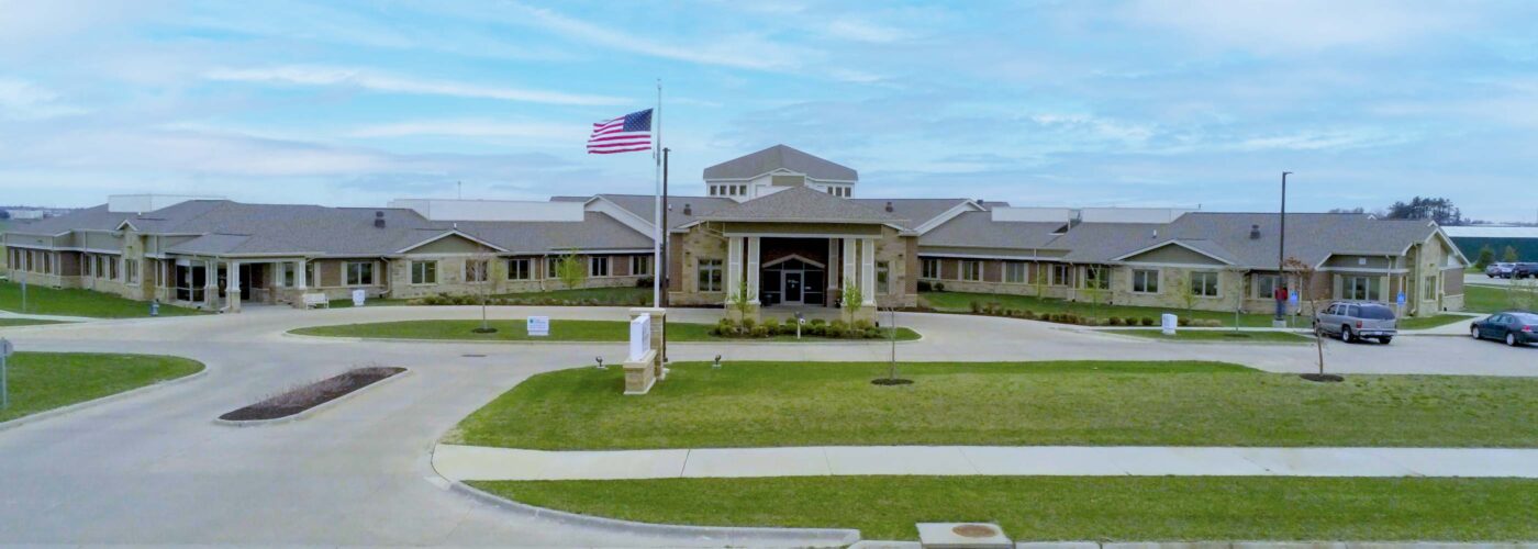 Under a blue sky, a large single-story building with a stone facade stands proudly. An American flag waves gently on the flagpole out front. This Care Initiative facility is surrounded by neatly maintained grass and a circular driveway, reflecting its welcoming atmosphere. Care Initiative embodies warmth and hospitality in this setting.