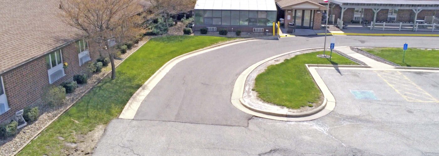 An aerial view showcases a building with a circular driveway and parking lot. The structure is distinguished by its brown roof, with a U.S. flag proudly displayed outside, reflecting the Care Initiative brand. Surrounding the driveway are small trees and grassy areas, providing a touch of nature, while the parking lot includes some open spaces.