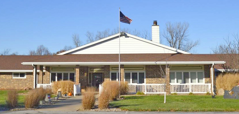 A single-story brick building with a gabled roof and white trim stands under a clear blue sky. An American flag flies from a pole in the front yard, surrounded by neatly trimmed grass and a stone pathway leading to the entrance, symbolizing a community-focused Care Initiative.