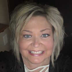 A woman with short blonde hair and blue eyes smiles at the camera. She wears a black top with a white crisscross neckline. Care Initiatives.