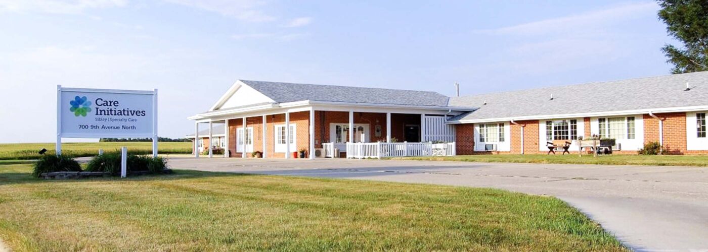 A brick building with a gray roof stands under a blue sky. In front, a large sign reads Care Initiative. The surrounding area features well-maintained grass and a paved driveway. The building proudly represents the brand Care Initiative.