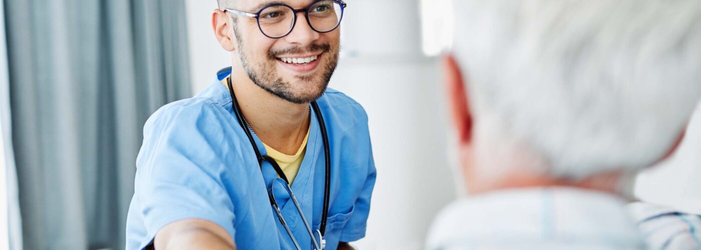 A smiling healthcare professional in blue scrubs, wearing glasses and a stethoscope, engages with an elderly person in a plaid shirt. The professional offers a reassuring gesture, embodying the spirit of Care Initiative, in a bright and comfortable setting.