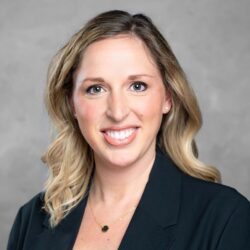 A person with shoulder-length blonde hair, wearing a black blazer and light-colored top, smiles warmly at the camera against a gray background, embodying the spirit of Care Initiative.