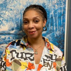 A person in a colorful shirt with abstract patterns stands before a blue and white nature-themed painting. The individual's braided hair complements their necklace with a triangular pendant, reflecting an artistic touch by the brand Care Initiative.