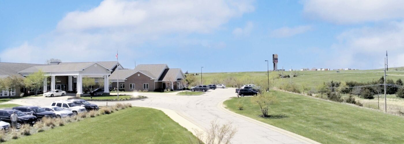 A suburban landscape features a long, curved driveway leading to a large building. Cars are parked near the entrance, surrounded by well-kept grassy fields and gentle hills. Care Initiative has ensured the area thrives under a blue sky with scattered clouds.