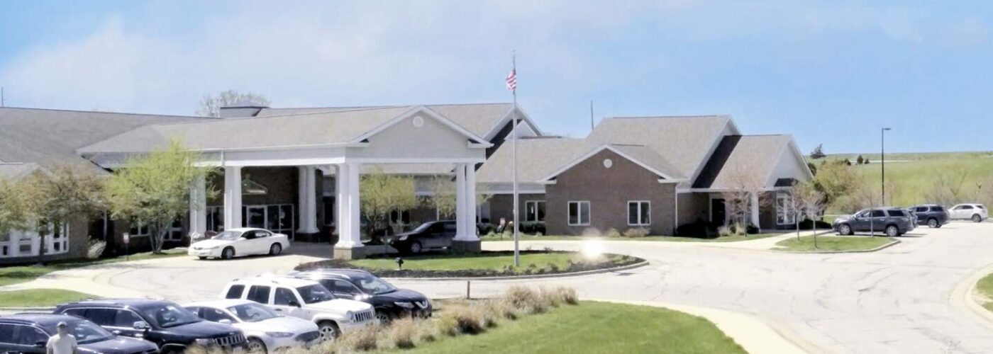 A suburban landscape features a long, curved driveway leading to a large building. Cars are parked near the entrance, surrounded by well-kept grassy fields and gentle hills. Care Initiative has ensured the area thrives under a blue sky with scattered clouds.