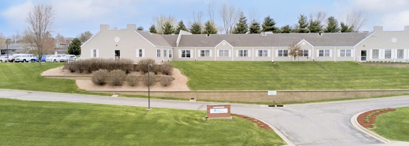 A large, single-story building with a beige exterior and gabled roof is situated on a grassy landscape. A wooden sign near the road highlights its role in the Care Initiative. Small trees and shrubs embellish the property under a partly cloudy sky.