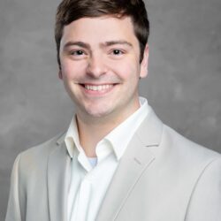 A man in a light gray suit and white shirt smiles warmly against a gray backdrop, embodying the spirit of Care Initiative.