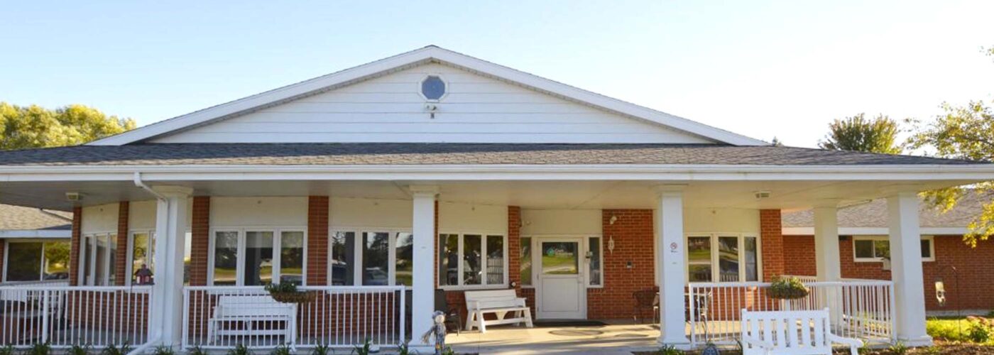 A single-story building with a brick facade and a white porch welcomes visitors with two benches flanking the entrance, while a concrete path leads the way. Enhanced by lush plants, this inviting space is perfect for pursuing Care Initiative under sunny, clear skies.