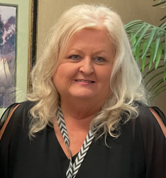 A woman with long white hair smiles in a black outfit paired with a chevron-patterned scarf. Behind her, a framed picture and green plant leaves can be seen, suggesting a calm environment, perhaps inspired by the Care Initiative.