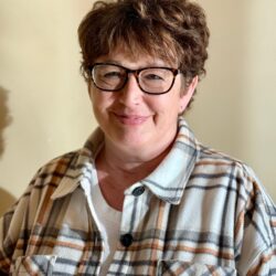 A person with short curly hair and glasses smiles warmly, wearing a plaid shirt in brown and grey over a white top. They're standing against a plain background with soft lighting, embodying the spirit of Care Initiative.