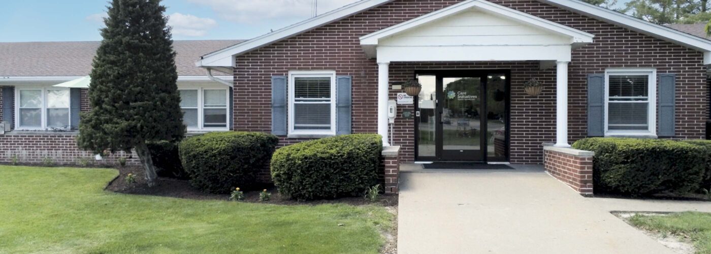 A brick building with a white portico and dark front doors is surrounded by neatly trimmed bushes and a lush green lawn, symbolizing the Care Initiative. A paved walkway leads to the entrance under a partly cloudy sky, representing Care Initiative.