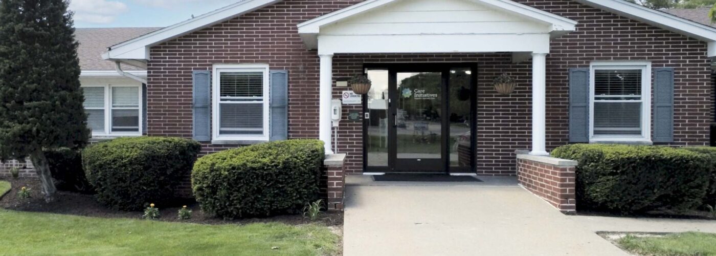 A brick building with a white portico and dark front doors is surrounded by neatly trimmed bushes and a lush green lawn, symbolizing the Care Initiative. A paved walkway leads to the entrance under a partly cloudy sky, representing Care Initiatives.