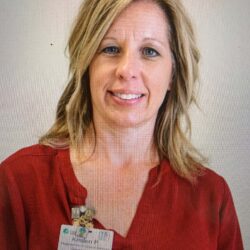 A woman with shoulder-length blonde hair, wearing a red blouse and name badge, smiles warmly against a plain background, embodying the spirit of Care Initiative.