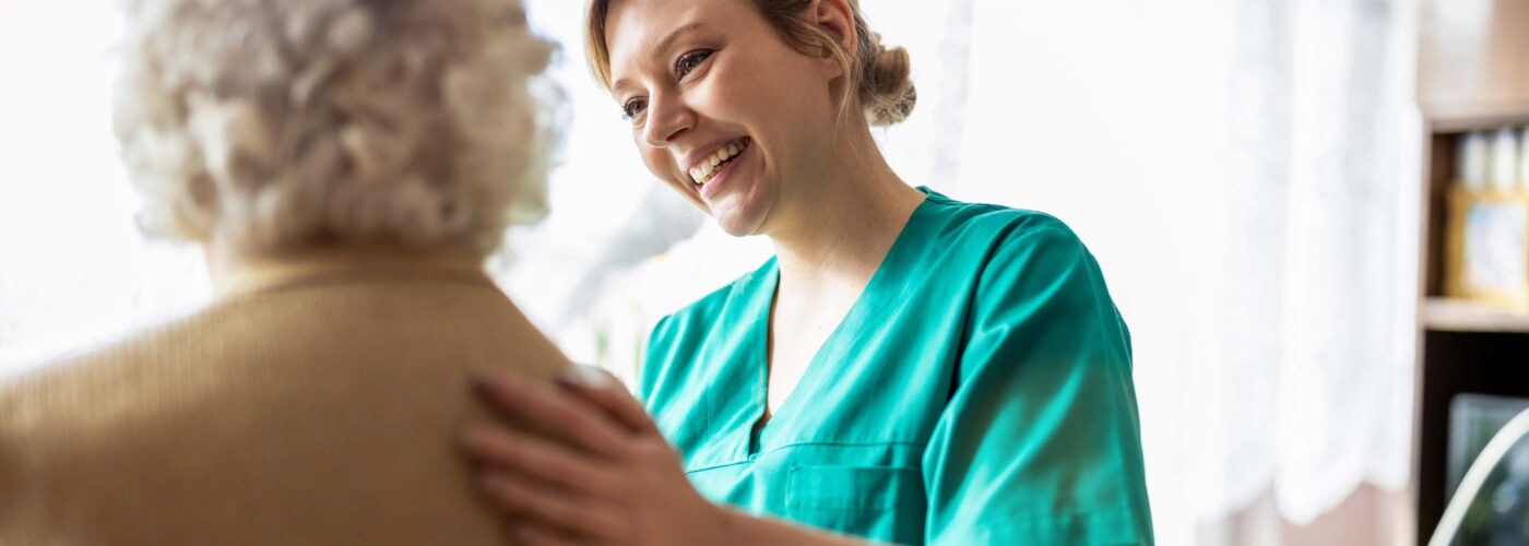 A caregiver in a green uniform is smiling and gently touching the shoulder of an elderly person with white hair, who is wearing a beige sweater. They are part of the Care Initiative in a bright room with large windows.