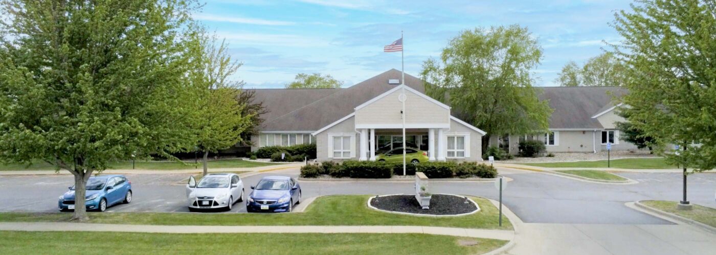 The single-story building with a pitched roof prominently features the American flag on a tall flagpole. Three cars are parked in front, while the area, surrounded by trees and grass under a blue sky with clouds, reflects the spirit of a community-driven Care Initiative. The brand name Care Initiative is central to this community-focused scene.