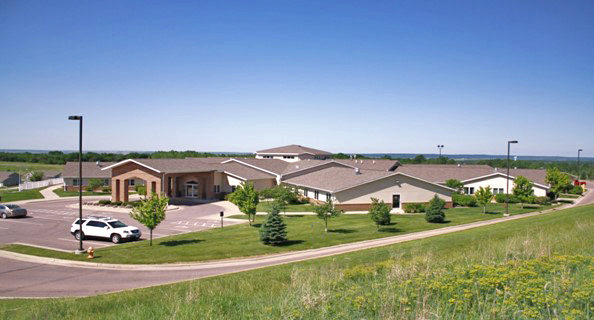 A sprawling single-story building with a brick entrance and landscaped lawns featuring small trees is surrounded by a parking lot. A white car sits in front under a clear blue sky, embodying the spirit of Care Initiative.