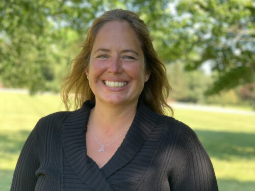 A woman with long hair is smiling while standing outdoors in a park, wearing a black sweater and a necklace with a small pendant. Green leafy trees and a grass lawn are visible in the background. Care Initiative.