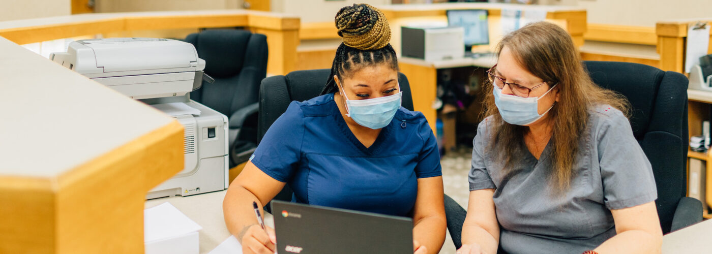 Two nurses wearing masks sit at a desk in a hospital, focused on reviewing papers and using a laptop. They collaborate efficiently as part of the Care Initiative. A printer and hallway are visible in the background, highlighting their dedicated work environment.