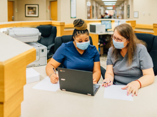 Two nurses wearing masks sit at a desk in a hospital, focused on reviewing papers and using a laptop. They collaborate efficiently as part of the Care Initiative. A printer and hallway are visible in the background, highlighting their dedicated work environment.