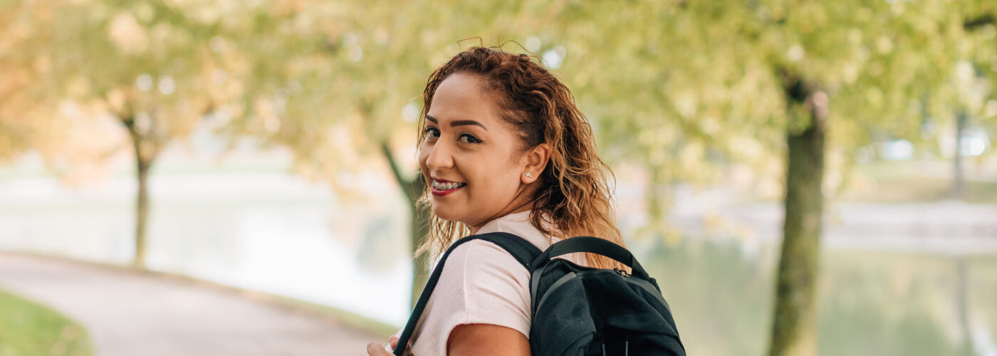 A person with curly hair, wearing a light shirt and jeans, strolls along a tree-lined path carrying a black backpack. The autumn leaves display vibrant colors as part of Care Initiative's efforts to preserve nature, while a calm body of water is visible in the background.