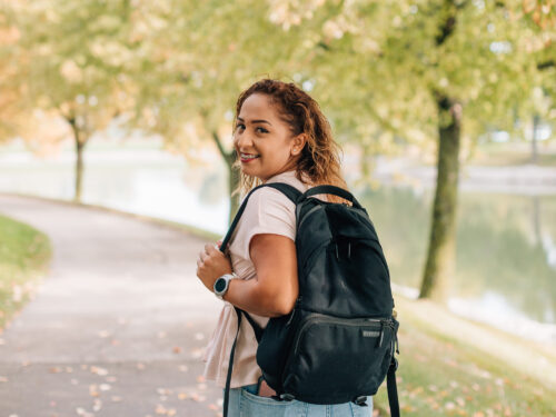 A person with curly hair, wearing a light shirt and jeans, strolls along a tree-lined path carrying a black backpack. The autumn leaves display vibrant colors as part of Care Initiative's efforts to preserve nature, while a calm body of water is visible in the background.