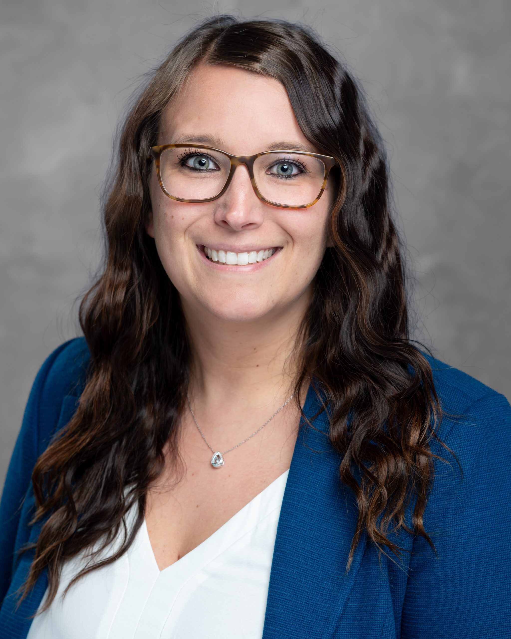 A woman with long wavy brown hair and glasses stands in a blue blazer over a white top, smiling warmly against a gray background. Her welcoming demeanor embodies the spirit of an inspiring Care Initiative.