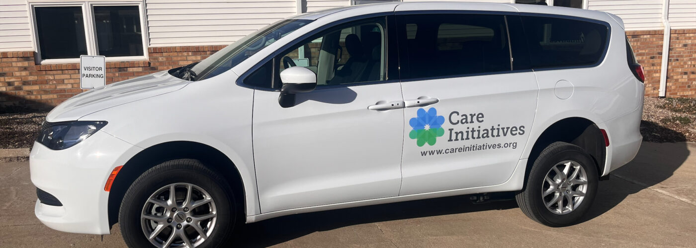 A white minivan featuring the Care Initiative logo and website is parked in front of a building that combines brick and white siding on a paved area. It's a sunny day with clear blue skies.