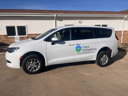 A white minivan featuring the Care Initiative logo and website is parked in front of a building that combines brick and white siding on a paved area. It's a sunny day with clear blue skies.