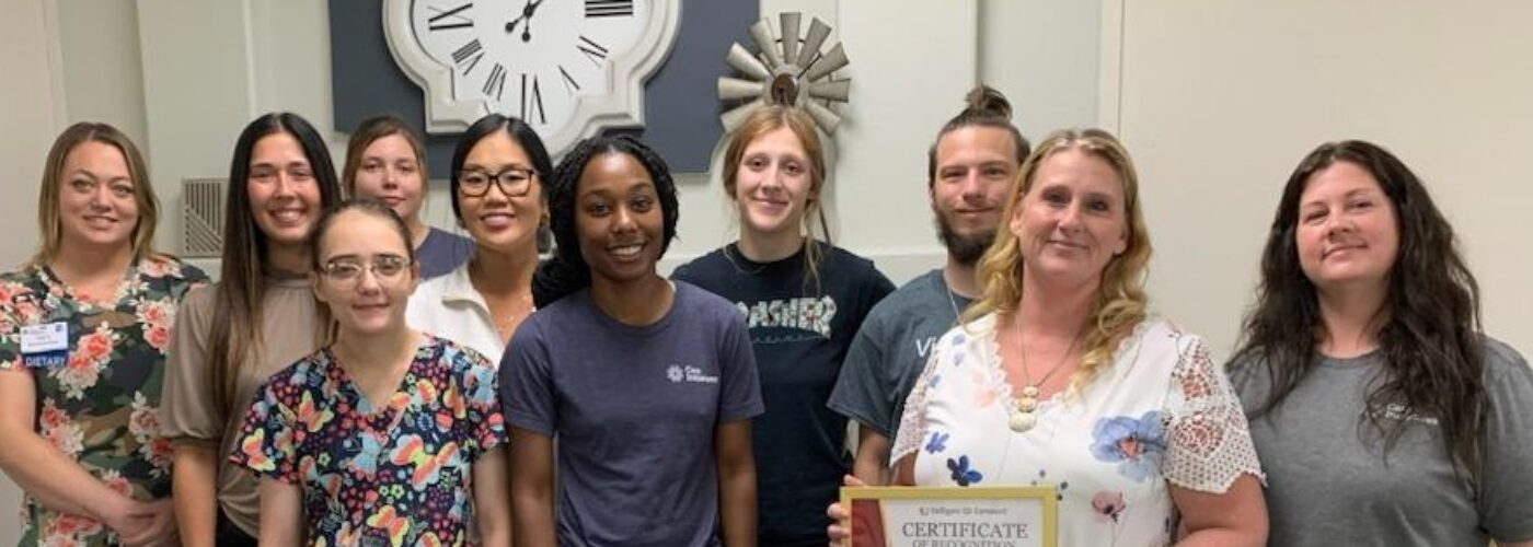 A group of ten people stands indoors, smiling at the camera as one proudly holds a framed certificate. A large wall clock is in the background, while they showcase casual and work attire. This gathering marks a significant moment for their Care Initiative.