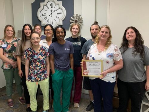 A group of ten people stands indoors, smiling at the camera as one proudly holds a framed certificate. A large wall clock is in the background, while they showcase casual and work attire. This gathering marks a significant moment for their Care Initiative.