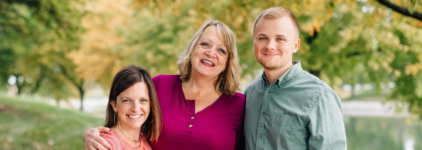 Three people are posing together outdoors in a park. Two women and one man stand before green trees and a path, smiling warmly. The woman in the center, embodying the spirit of Care Initiative, has her arms around the others.